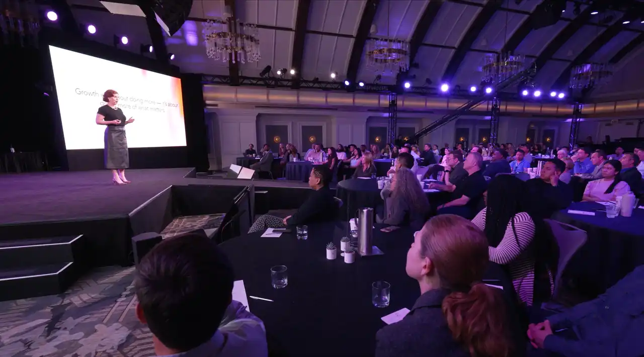 A packed hotel ballroom watching a keynote from round tables under chandeliers